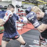 Two fighters sparring in a gym, wearing gloves and training gear inside a boxing ring.
