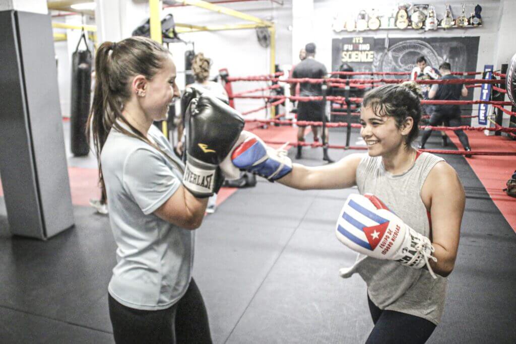 Two women in boxing gloves smiling while sparring in a gym ring.