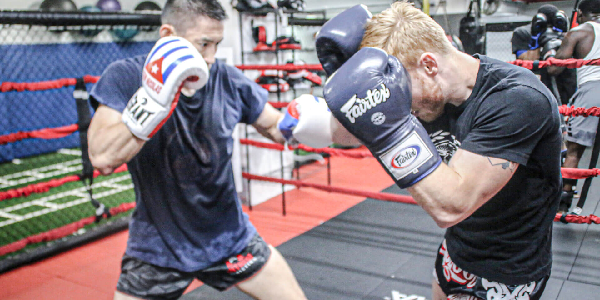 Two fighters sparring in a gym, wearing gloves and training gear inside a boxing ring.